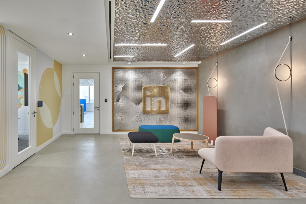 Modern office reception area with contemporary furniture including a pale pink sofa, black and green benches, and a round coffee table on a patterned rug. The backdrop features a decorative metallic LinkedIn logo wall with a textured silver finish. The ceiling has artistic metallic panels with integrated linear lights. The space also includes artistic wall decor and minimalistic pendant lighting.