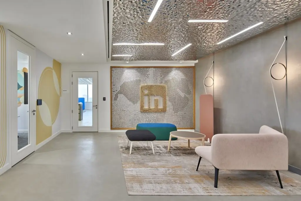 Modern office reception area with contemporary furniture including a pale pink sofa, black and green benches, and a round coffee table on a patterned rug. The backdrop features a decorative metallic LinkedIn logo wall with a textured silver finish. The ceiling has artistic metallic panels with integrated linear lights. The space also includes artistic wall decor and minimalistic pendant lighting.