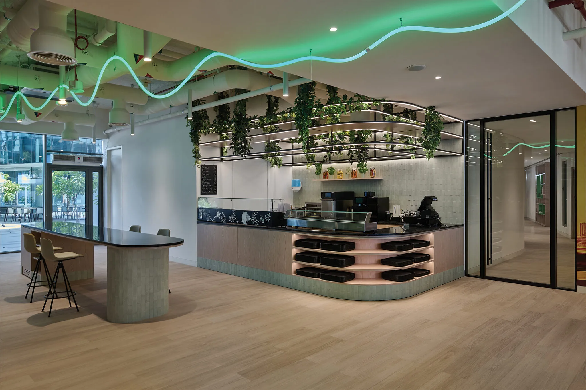 Pantry area from Schneider Electric’s sustainable office fit-out in Dubai, featuring a green wave ceiling light, hanging plants, timber finishes, and modern counter seating